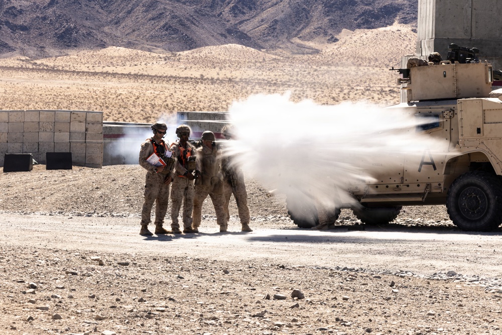 Military personnel in desert camouflage uniforms with orange observer/safety vests stand near a tan armored military vehicle (likely an MRAP or HMMWV) during what appears to be a live-fire training exercise. A projectile or blast effect is visible emanating from the vehicle's side, creating a white smoke/dust cloud. The setting is an arid desert training range with rocky terrain and mountains in the background.