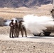 Military personnel in desert camouflage uniforms with orange observer/safety vests stand near a tan armored military vehicle (likely an MRAP or HMMWV) during what appears to be a live-fire training exercise. A projectile or blast effect is visible emanating from the vehicle's side, creating a white smoke/dust cloud. The setting is an arid desert training range with rocky terrain and mountains in the background.