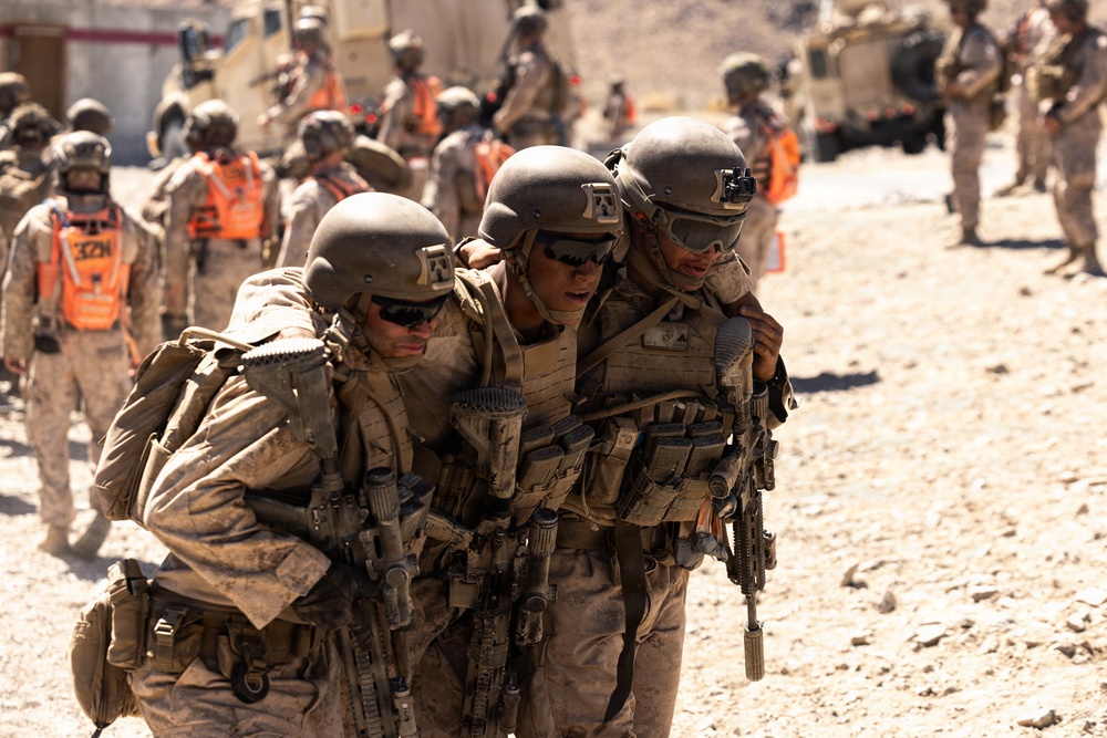 Military personnel in desert camouflage uniforms are conducting what appears to be a casualty evacuation drill or training exercise in an arid environment. Two soldiers are supporting and carrying a third soldier between them in a buddy-carry technique. Background personnel wearing bright orange safety vests indicate this is a supervised training exercise rather than a combat situation.