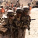 Military personnel in desert camouflage uniforms are conducting what appears to be a casualty evacuation drill or training exercise in an arid environment. Two soldiers are supporting and carrying a third soldier between them in a buddy-carry technique. Background personnel wearing bright orange safety vests indicate this is a supervised training exercise rather than a combat situation.