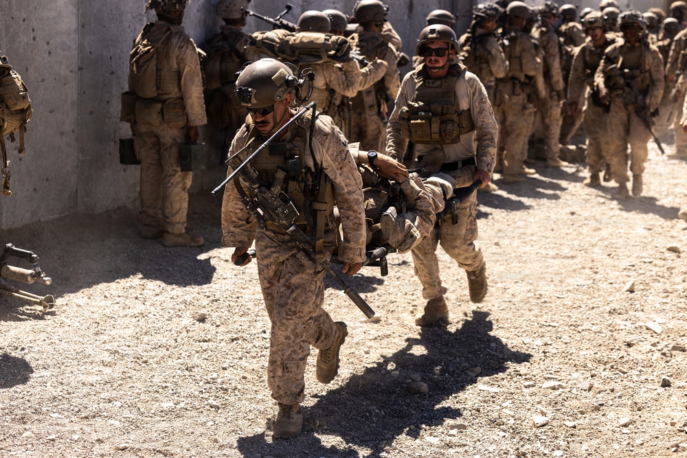 The image shows a group of military personnel in desert camouflage uniforms conducting what appears to be a tactical operation or training exercise. Two soldiers in the foreground are carrying what appears to be a casualty or simulated casualty on a stretcher while moving urgently. A larger formation of similarly equipped soldiers is visible in the background near a concrete wall structure.