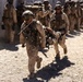 The image shows a group of military personnel in desert camouflage uniforms conducting what appears to be a tactical operation or training exercise. Two soldiers in the foreground are carrying what appears to be a casualty or simulated casualty on a stretcher while moving urgently. A larger formation of similarly equipped soldiers is visible in the background near a concrete wall structure.