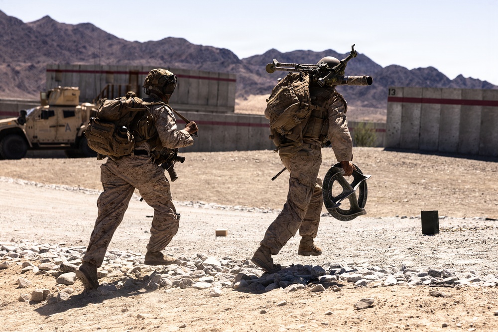 Two military personnel in desert camouflage uniforms are moving rapidly across a rocky, arid training area. One soldier carries a large crew-served weapon system on their back while holding what appears to be an ammunition drum. The setting appears to be a military training range, evidenced by concrete barriers, a tactical vehicle, and numbered range markers in the background.