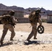 Two military personnel in desert camouflage uniforms are moving rapidly across a rocky, arid training area. One soldier carries a large crew-served weapon system on their back while holding what appears to be an ammunition drum. The setting appears to be a military training range, evidenced by concrete barriers, a tactical vehicle, and numbered range markers in the background.