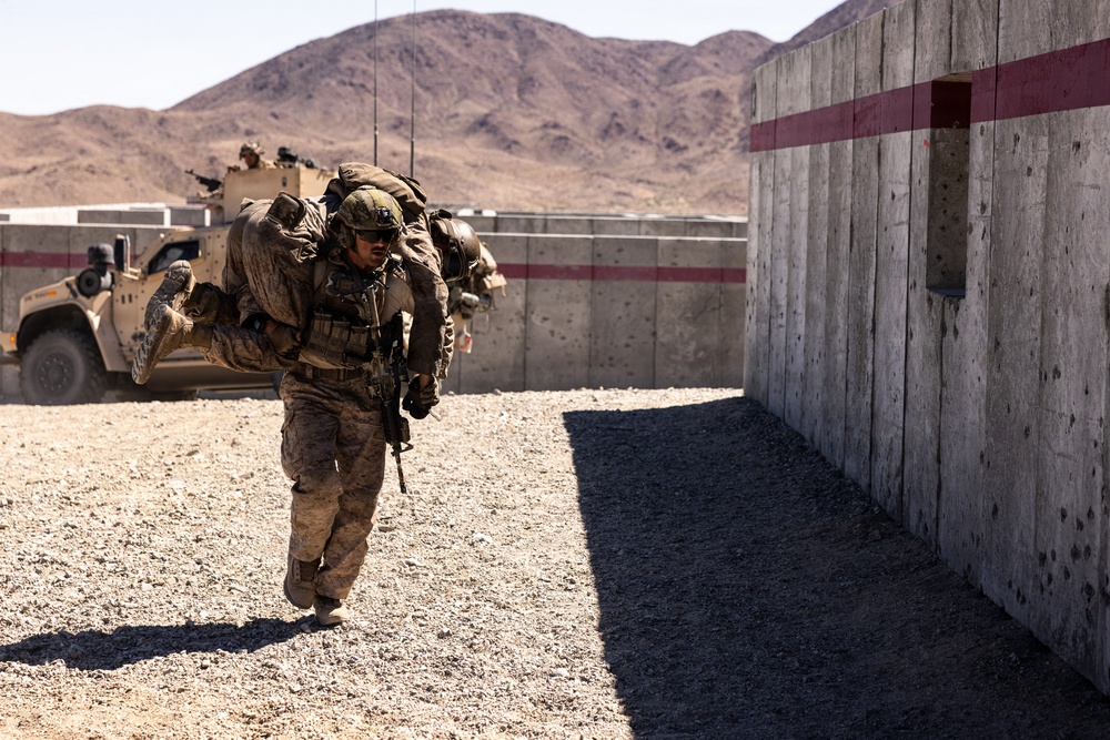 A military service member in desert camouflage (MARPAT pattern) is carrying what appears to be a simulated casualty over their shoulders in a fireman's carry position. The scene takes place in an arid, desert training environment with concrete barrier walls marked with red stripes and a tactical military vehicle (JLTV or similar MRAP-class vehicle) visible in the background. This appears to be a combat casualty evacuation (CASEVAC) training exercise.
