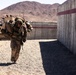 A military service member in desert camouflage (MARPAT pattern) is carrying what appears to be a simulated casualty over their shoulders in a fireman's carry position. The scene takes place in an arid, desert training environment with concrete barrier walls marked with red stripes and a tactical military vehicle (JLTV or similar MRAP-class vehicle) visible in the background. This appears to be a combat casualty evacuation (CASEVAC) training exercise.