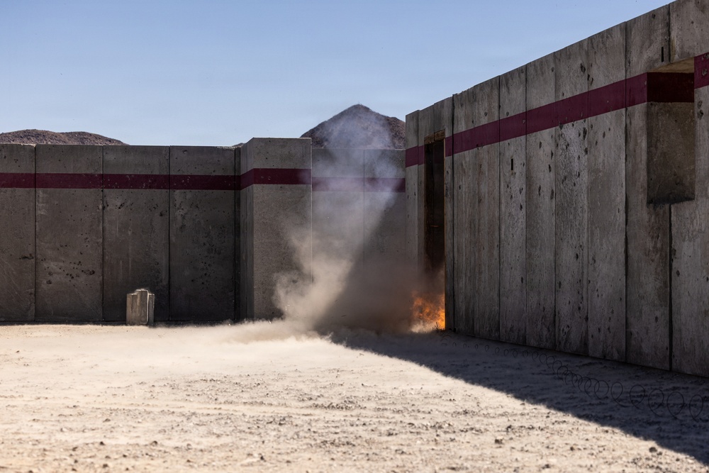 The image shows a controlled explosive or breaching event occurring at a concrete training facility in an arid desert environment. A plume of dust and smoke rises from a doorway or gap between concrete wall sections, accompanied by visible orange flames at the base. The facility appears to be a military or law enforcement training range with concrete blast walls marked with dark red horizontal stripes.