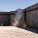 The image shows a controlled explosive or breaching event occurring at a concrete training facility in an arid desert environment. A plume of dust and smoke rises from a doorway or gap between concrete wall sections, accompanied by visible orange flames at the base. The facility appears to be a military or law enforcement training range with concrete blast walls marked with dark red horizontal stripes.