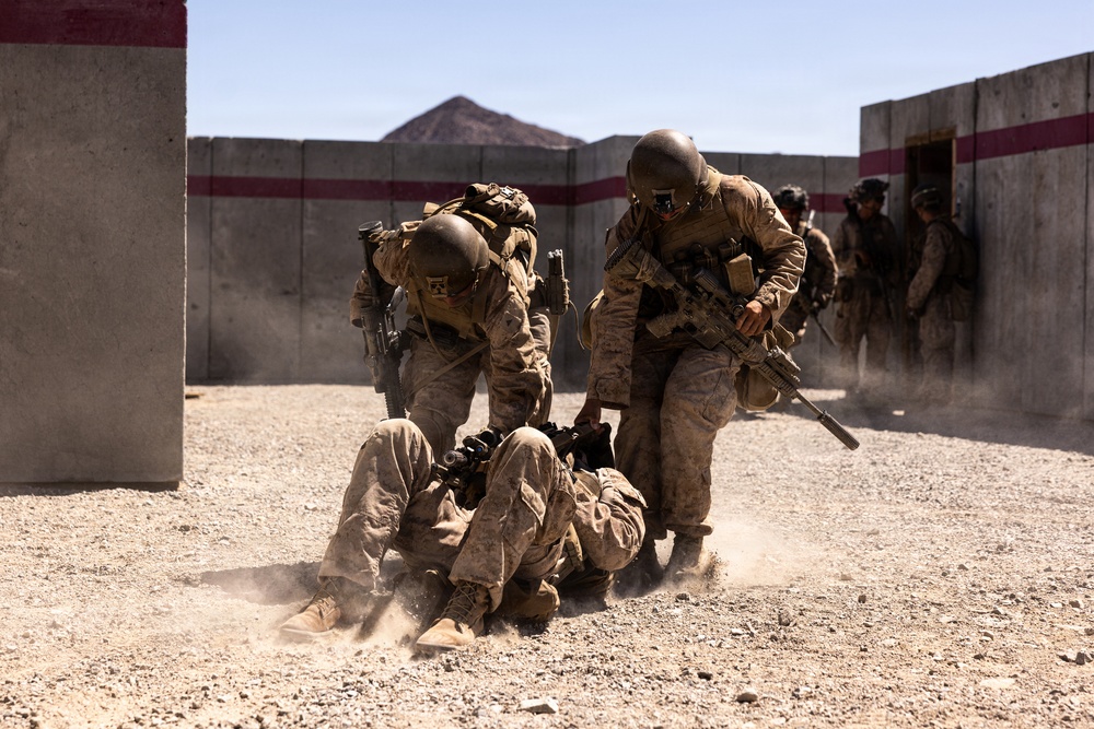 Military personnel in desert camouflage uniforms are conducting what appears to be a training exercise at a mock urban environment or combat training facility. Several soldiers are assisting or extracting a downed personnel from the ground while others stand by in the background. The setting features concrete walls with red and white markings typical of military training ranges.