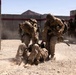 Military personnel in desert camouflage uniforms are conducting what appears to be a training exercise at a mock urban environment or combat training facility. Several soldiers are assisting or extracting a downed personnel from the ground while others stand by in the background. The setting features concrete walls with red and white markings typical of military training ranges.
