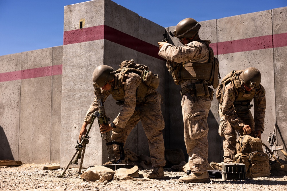 Three military personnel in desert camouflage uniforms and tactical gear are positioned against a concrete wall with red horizontal stripes, appearing to be engaged in a training exercise. One soldier is handling a weapon with a bipod on the ground, another is observing around the corner of the wall, and a third is accessing equipment from a pack. The arid environment, concrete barrier structure, and organized gear suggest a controlled military training facility.