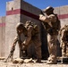 Three military personnel in desert camouflage uniforms and tactical gear are positioned against a concrete wall with red horizontal stripes, appearing to be engaged in a training exercise. One soldier is handling a weapon with a bipod on the ground, another is observing around the corner of the wall, and a third is accessing equipment from a pack. The arid environment, concrete barrier structure, and organized gear suggest a controlled military training facility.