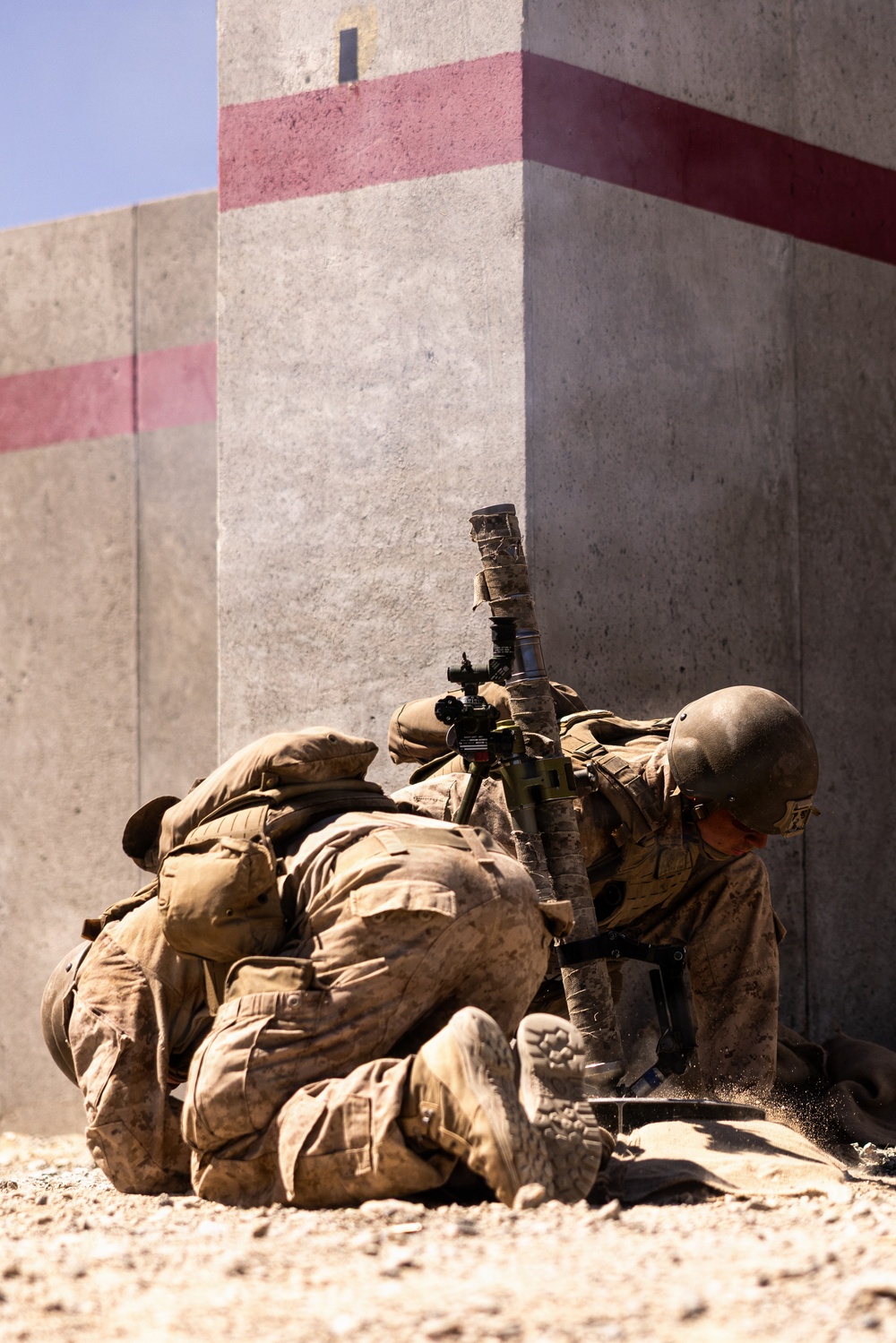 Two military personnel in desert camouflage uniforms are positioned at the base of a concrete wall corner, taking cover in a low prone/crouching position. One soldier is equipped with a weapon system featuring a suppressor, optic sight, and bipod. The setting appears to be a military training environment or urban operations area, identifiable by the concrete barriers marked with red/pink painted stripes.