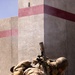Two military personnel in desert camouflage uniforms are positioned at the base of a concrete wall corner, taking cover in a low prone/crouching position. One soldier is equipped with a weapon system featuring a suppressor, optic sight, and bipod. The setting appears to be a military training environment or urban operations area, identifiable by the concrete barriers marked with red/pink painted stripes.