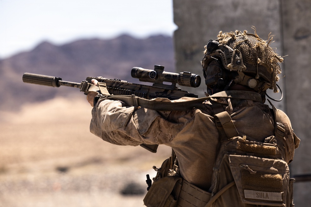 A military service member in full combat gear is aiming a scoped rifle with a suppressor attached in an arid, desert environment. The individual is wearing MARPAT (Marine pattern) camouflage uniform with tactical equipment and a ghillie-style helmet cover. A name tape reading 'SULA' is visible on the back of their gear.