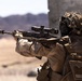 A military service member in full combat gear is aiming a scoped rifle with a suppressor attached in an arid, desert environment. The individual is wearing MARPAT (Marine pattern) camouflage uniform with tactical equipment and a ghillie-style helmet cover. A name tape reading 'SULA' is visible on the back of their gear.