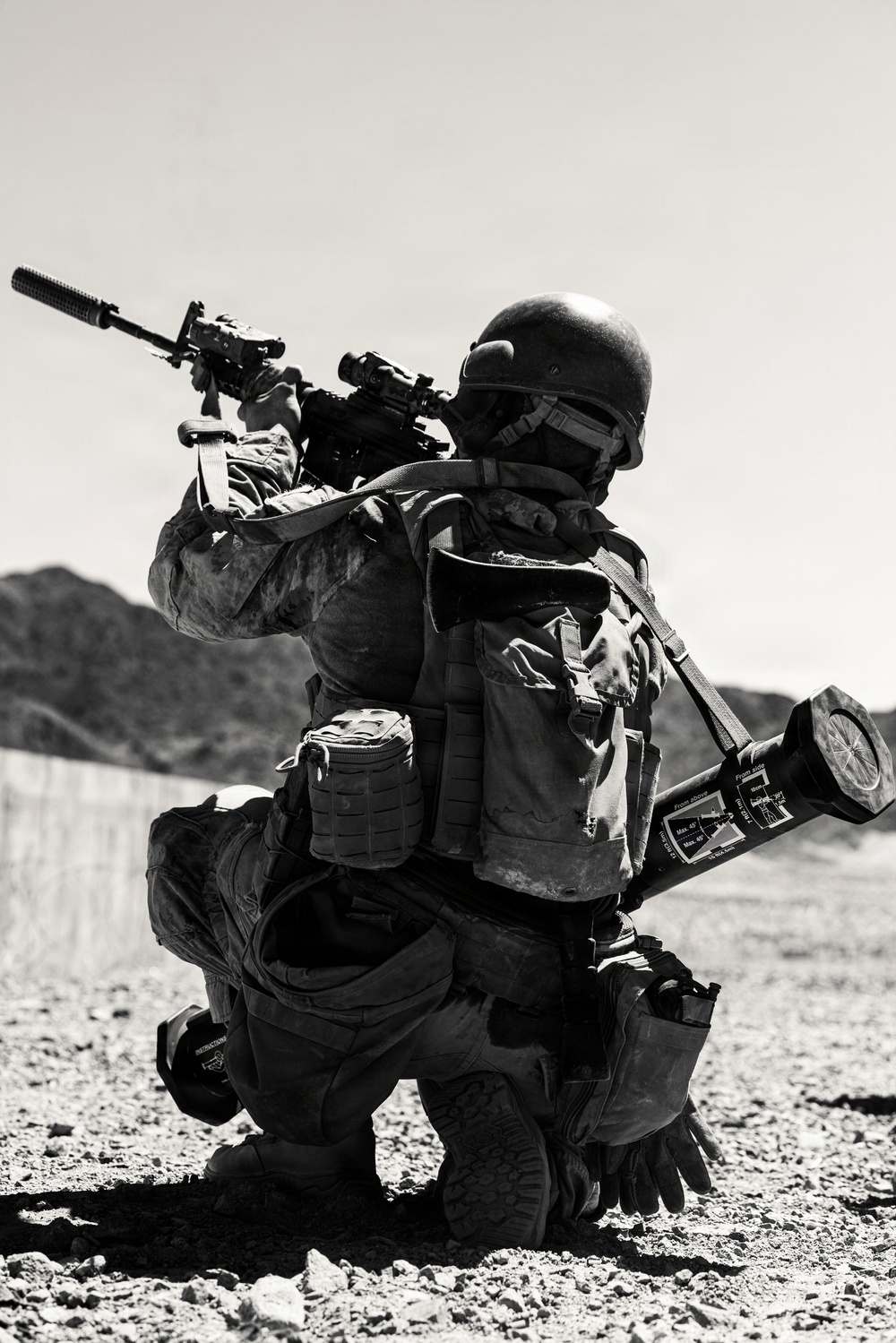 A heavily equipped soldier is kneeling on rocky desert terrain in a firing position, aiming a suppressed rifle with an optic sight. The soldier carries extensive gear including a tactical vest, backpack, and what appears to be a rocket launcher or anti-tank weapon slung across the back. The black-and-white photograph is taken in an arid, desert-like environment with hills visible in the background.