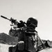 A heavily equipped soldier is kneeling on rocky desert terrain in a firing position, aiming a suppressed rifle with an optic sight. The soldier carries extensive gear including a tactical vest, backpack, and what appears to be a rocket launcher or anti-tank weapon slung across the back. The black-and-white photograph is taken in an arid, desert-like environment with hills visible in the background.