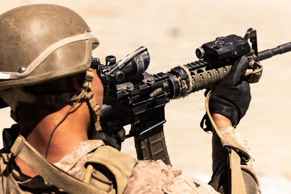 A soldier in desert camouflage uniform and tactical helmet is aiming a rifle equipped with multiple optical attachments. The image is taken from a rear-angle perspective in what appears to be an arid, sandy environment. The soldier is in a firing stance with both hands gripping the weapon.