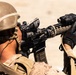 A soldier in desert camouflage uniform and tactical helmet is aiming a rifle equipped with multiple optical attachments. The image is taken from a rear-angle perspective in what appears to be an arid, sandy environment. The soldier is in a firing stance with both hands gripping the weapon.