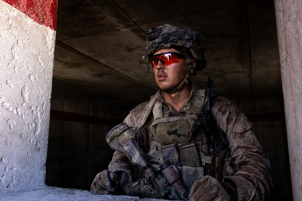 A soldier in full combat gear is positioned in a covered concrete structure, appearing to be in an alert or observational stance. The individual is equipped with camouflage uniform, tactical vest, helmet with netting, and distinctive red-tinted ballistic eyewear. The setting suggests an urban operations environment, possibly during a training exercise or active deployment.