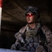A soldier in full combat gear is positioned in a covered concrete structure, appearing to be in an alert or observational stance. The individual is equipped with camouflage uniform, tactical vest, helmet with netting, and distinctive red-tinted ballistic eyewear. The setting suggests an urban operations environment, possibly during a training exercise or active deployment.