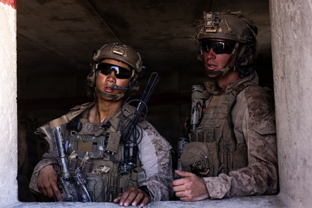 Two armed military personnel in full combat gear are positioned at what appears to be a window or opening in a concrete structure. Both wear MARPAT (Marine pattern) camouflage uniforms, tactical vests, helmets with accessories, and ballistic sunglasses, suggesting they are U.S. Marines. They appear to be conducting an observation or security posture during an operation or training exercise.