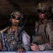 Two armed military personnel in full combat gear are positioned at what appears to be a window or opening in a concrete structure. Both wear MARPAT (Marine pattern) camouflage uniforms, tactical vests, helmets with accessories, and ballistic sunglasses, suggesting they are U.S. Marines. They appear to be conducting an observation or security posture during an operation or training exercise.