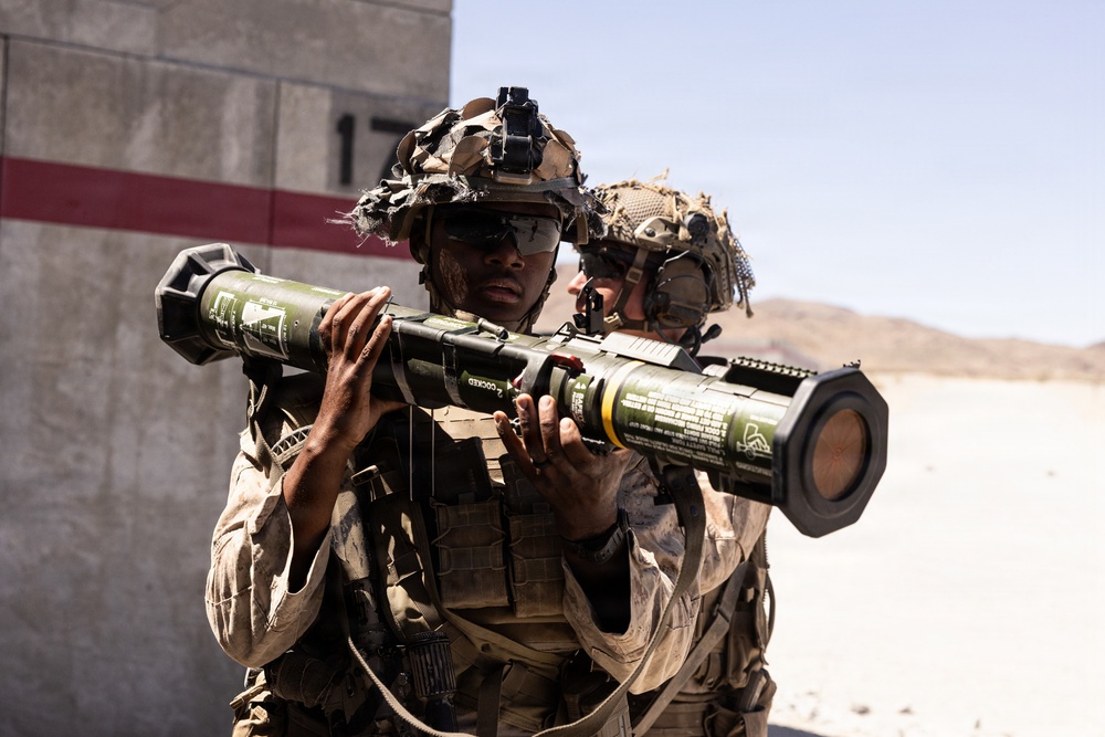 Two military personnel in desert camouflage uniforms and tactical gear are handling a shoulder-fired rocket launcher, likely an AT4 or similar anti-armor weapon system. The setting appears to be a desert training range, indicated by the arid terrain, numbered range marker visible in the background, and the overall training context. Both soldiers are wearing combat helmets with accessories and tactical eye protection.