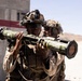 Two military personnel in desert camouflage uniforms and tactical gear are handling a shoulder-fired rocket launcher, likely an AT4 or similar anti-armor weapon system. The setting appears to be a desert training range, indicated by the arid terrain, numbered range marker visible in the background, and the overall training context. Both soldiers are wearing combat helmets with accessories and tactical eye protection.