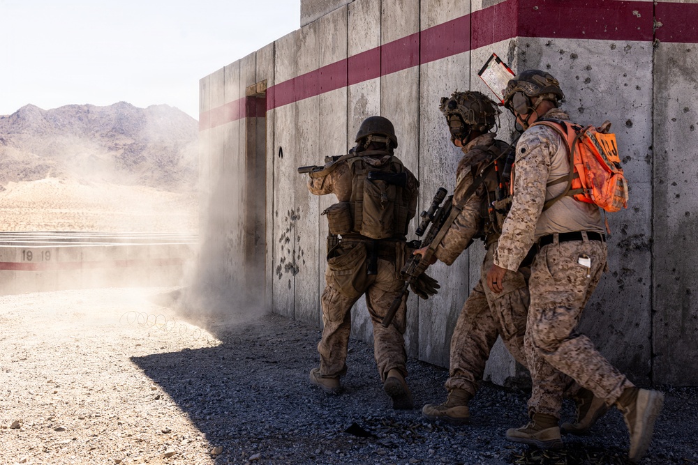 Three military personnel in desert camouflage uniforms are conducting a tactical training exercise at what appears to be a military training range in an arid environment. The soldiers are moving along a concrete wall with bullet impact marks, with one soldier aiming a pistol around a corner. One individual is wearing a bright orange safety vest, indicating an observer or safety officer role during the exercise.