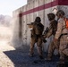 Three military personnel in desert camouflage uniforms are conducting a tactical training exercise at what appears to be a military training range in an arid environment. The soldiers are moving along a concrete wall with bullet impact marks, with one soldier aiming a pistol around a corner. One individual is wearing a bright orange safety vest, indicating an observer or safety officer role during the exercise.