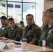 A group of Brazilian military personnel are seated at a conference table during what appears to be a formal briefing or multinational military meeting. Several individuals are wearing green flight suits and camouflage uniforms with 'BRASIL' patches and name badges. The setting appears to be a modern conference or meeting facility with large windows.