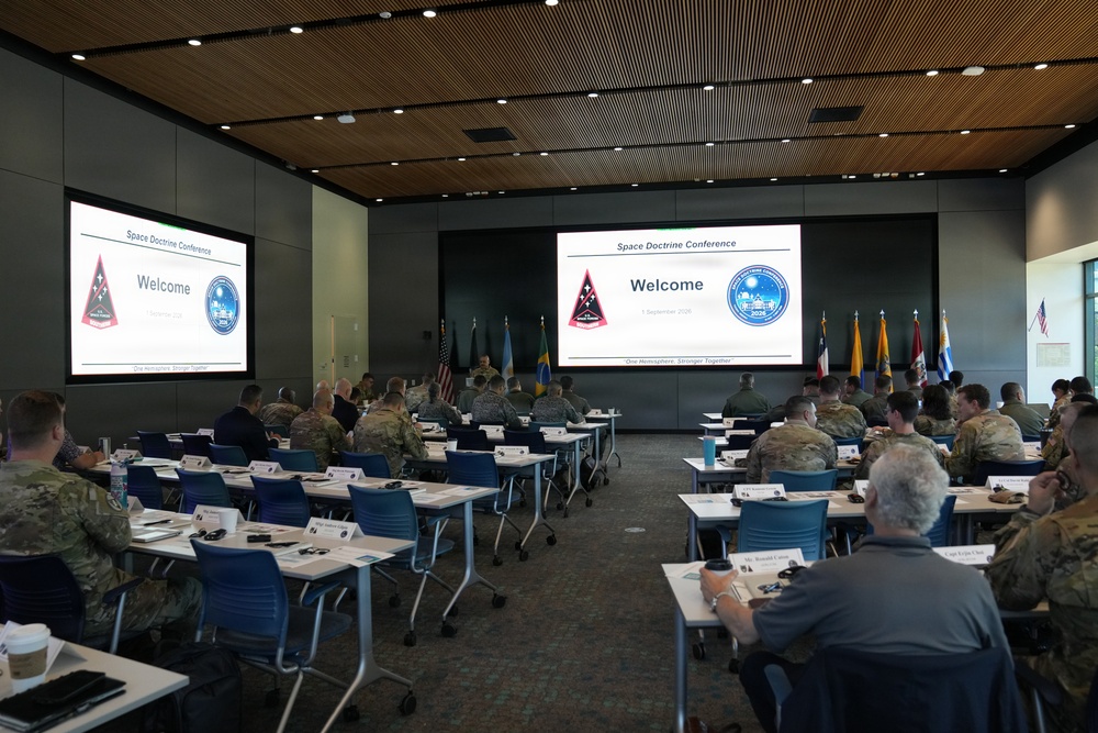 A military conference room is set up for a 'Space Doctrine Conference' with attendees seated at tables facing two large projection screens displaying a 'Welcome' slide. The majority of attendees appear to be military personnel in camouflage uniforms, with a few civilians present. Multiple national and organizational flags are displayed at the front of the room.