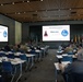 A military conference room is set up for a 'Space Doctrine Conference' with attendees seated at tables facing two large projection screens displaying a 'Welcome' slide. The majority of attendees appear to be military personnel in camouflage uniforms, with a few civilians present. Multiple national and organizational flags are displayed at the front of the room.