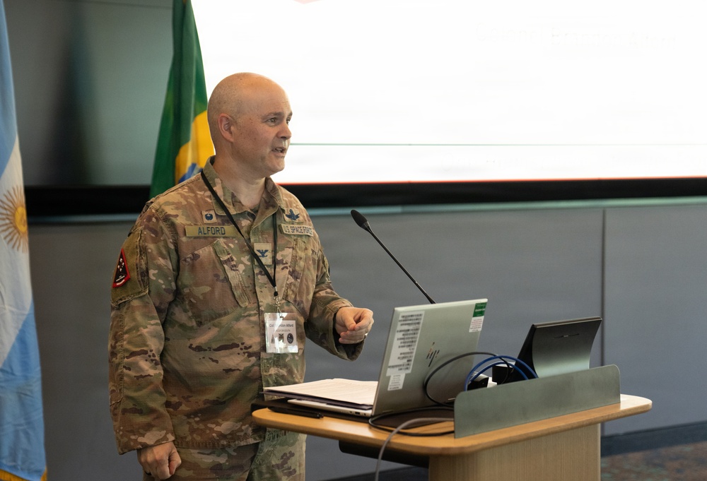 A U.S. Space Force officer identified by name tape as 'ALFORD' is delivering a presentation at a podium equipped with a laptop and microphone. The setting appears to be an international military or diplomatic conference, as evidenced by the Argentine and Brazilian flags visible in the background. The officer wears the OCP (Operational Camouflage Pattern) uniform with a colonel's rank insignia (eagle) and a conference lanyard/badge.