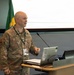 A U.S. Space Force officer identified by name tape as 'ALFORD' is delivering a presentation at a podium equipped with a laptop and microphone. The setting appears to be an international military or diplomatic conference, as evidenced by the Argentine and Brazilian flags visible in the background. The officer wears the OCP (Operational Camouflage Pattern) uniform with a colonel's rank insignia (eagle) and a conference lanyard/badge.