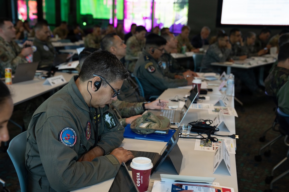 A military conference or briefing session with numerous personnel in various military uniforms seated at long tables with laptops and documents. The foreground shows a man in an olive flight suit with patches, wearing glasses and an earpiece, focused on a laptop. A large projection screen is visible in the background.