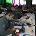 A military conference or briefing session with numerous personnel in various military uniforms seated at long tables with laptops and documents. The foreground shows a man in an olive flight suit with patches, wearing glasses and an earpiece, focused on a laptop. A large projection screen is visible in the background.