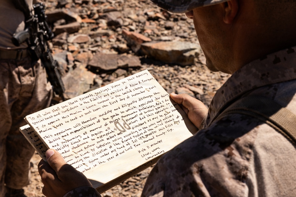A military service member in desert camouflage uniform is reading a handwritten document, appearing to be a promotion certificate or appointment letter. The document references a Lance Corporal promotion in the United States Marine Corps, signed by 'Kyle P.' with the title 'LtCol, USMC Commanding'. The setting appears to be an arid, rocky outdoor environment.