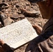 A military service member in desert camouflage uniform is reading a handwritten document, appearing to be a promotion certificate or appointment letter. The document references a Lance Corporal promotion in the United States Marine Corps, signed by 'Kyle P.' with the title 'LtCol, USMC Commanding'. The setting appears to be an arid, rocky outdoor environment.