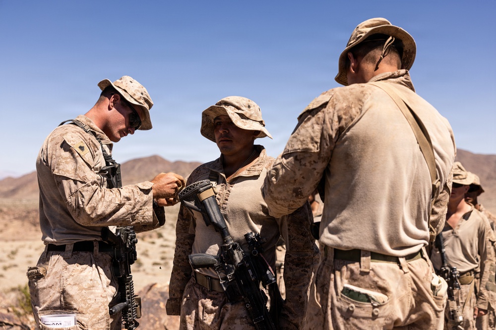 Military personnel in desert camouflage uniforms are gathered in an arid, desert environment. Two soldiers appear to be assisting or inspecting a third soldier's equipment, specifically handling what appears to be a weapon or gear attachment. The setting suggests a field training exercise or operational deployment in a desert region.