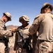 Military personnel in desert camouflage uniforms are gathered in an arid, desert environment. Two soldiers appear to be assisting or inspecting a third soldier's equipment, specifically handling what appears to be a weapon or gear attachment. The setting suggests a field training exercise or operational deployment in a desert region.