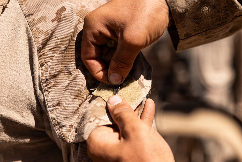 A close-up photograph showing two sets of hands working with military desert camouflage uniforms. One person appears to be pinning or attaching a small metallic item, possibly a rank insignia or pin, onto a Velcro patch on the sleeve or collar area of a MARPAT desert camouflage uniform. The action suggests a formal or ceremonial pinning event.