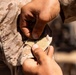 A close-up photograph showing two sets of hands working with military desert camouflage uniforms. One person appears to be pinning or attaching a small metallic item, possibly a rank insignia or pin, onto a Velcro patch on the sleeve or collar area of a MARPAT desert camouflage uniform. The action suggests a formal or ceremonial pinning event.