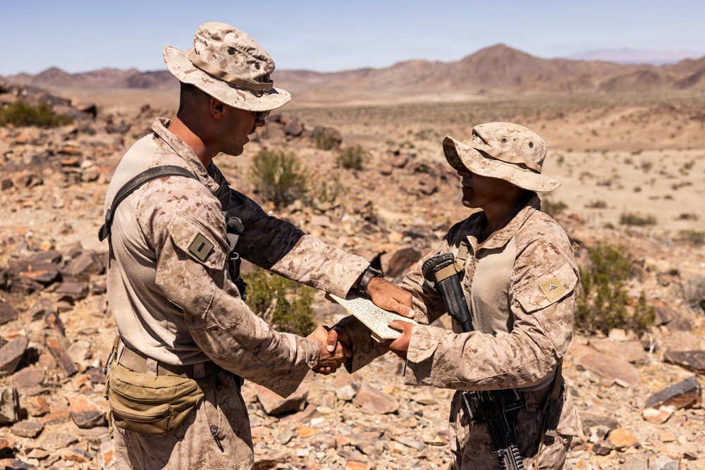 Two military personnel in desert camouflage uniforms are shaking hands while exchanging a document in an arid, rocky desert environment. The individual on the left appears to be an officer, identifiable by a rank insignia visible on the chest. The setting suggests a field training exercise or operational briefing in a desert location.