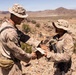 Two military personnel in desert camouflage uniforms are shaking hands while exchanging a document in an arid, rocky desert environment. The individual on the left appears to be an officer, identifiable by a rank insignia visible on the chest. The setting suggests a field training exercise or operational briefing in a desert location.