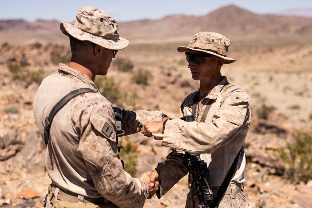 Two military personnel in desert camouflage uniforms are engaged in what appears to be a medal or coin presentation ceremony in an arid desert environment. One individual is pinning or presenting something to the other while simultaneously shaking hands, a common military recognition ritual. The setting appears to be an active training or operational desert location based on the terrain and equipment visible.
