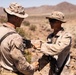 Two military personnel in desert camouflage uniforms are engaged in what appears to be a medal or coin presentation ceremony in an arid desert environment. One individual is pinning or presenting something to the other while simultaneously shaking hands, a common military recognition ritual. The setting appears to be an active training or operational desert location based on the terrain and equipment visible.