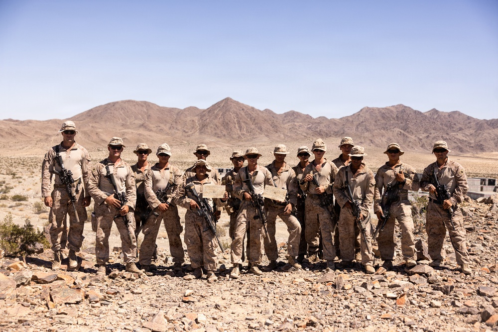 A group of approximately 13 military personnel in desert camouflage uniforms pose for a group photograph in an arid desert environment. They are armed with rifles and wearing patrol caps, standing on rocky terrain with barren mountains visible in the background. The setting and uniforms are consistent with a U.S. Marine Corps training exercise or operational deployment in a desert region.